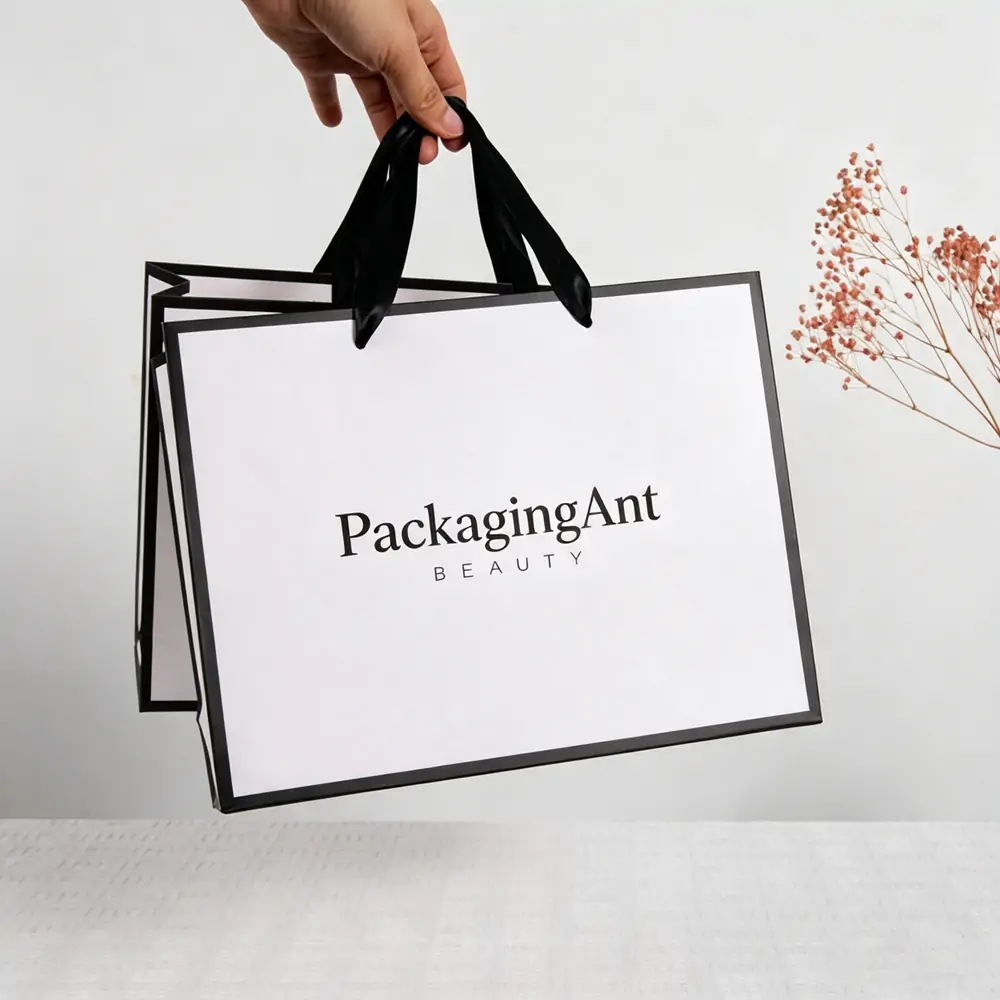 Hand holding a white luxury paper shopping bag with stylish black border trim and matching black ribbon handles.